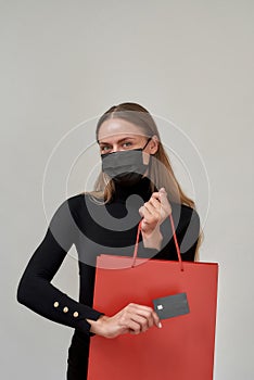 Pretty young woman wearing black protective mask holding plastic credit card while posing with red shopping bag isolated