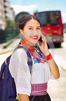 Pretty young woman wearing traditional andean blouse and blue backpack, waiting for bus at outdoors station platform