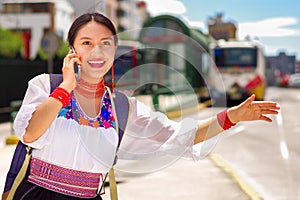 Pretty young woman wearing traditional andean blouse and blue backpack, waiting for bus at outdoors station platform