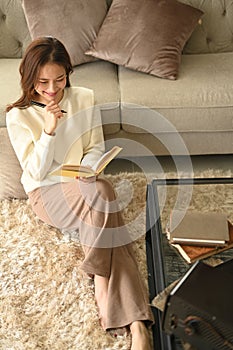 Pretty young woman wearing warm sweater sitting on carpet and reading book. Winter, coziness, leisure and people concept