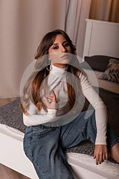 Pretty young woman in a white top in stylish jeans posing on a bed in the bedroom with a modern interior. Model of a trendy