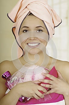 Pretty young woman wrapped with bath towels holding shampoo and loofah sponge