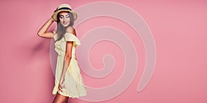 Pretty young woman in yellow dress and straw hat posing on pink background