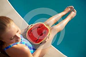 Prety blonde girl holding half of watermelon in the blue pool on slim legs. Young woman wearing blue stripedbody