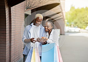 Prices and sales monitoring. African american millennial couple with colors bags looking at smartphone