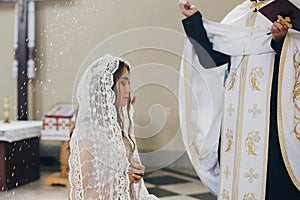 Priest blessing with holy water stylish bride in kerchief at altar during holy matrimony in church. Wedding ceremony in cathedral