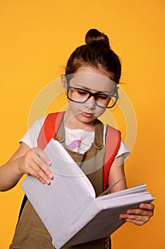 Primary school girl in casual wear and glasses, learns reading alphabet, holding mockup book, isolated orange background