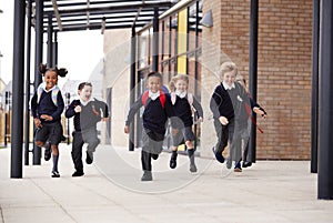 Primary school kids, wearing school uniforms and backpacks, running on a walkway outside their school building, front view