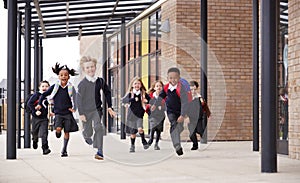 Primary school kids, wearing school uniforms and backpacks, running on a walkway outside their school building, front view