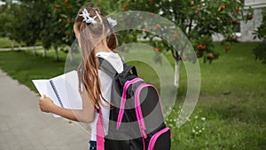 Primary school student with a book in his hands. Girl with a backpack near the school. Beginning of lessons. First day of autumn.