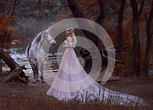 The princess met a unicorn in the forest. The blonde girl with a gentle make-up, is dressed in a long vintage dress with