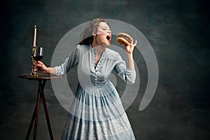 Princess. Portrait of aristocratic woman wearing blue historical dress eating big hamburger. Contemporary food art.