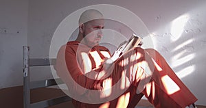 prisoner in orange overalls reading a book in a prison cell sitting on the bed.