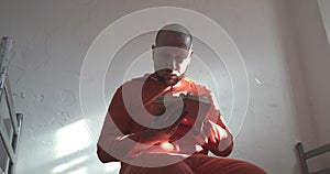 prisoner in orange overalls reading a book in a prison cell sitting on the bed.