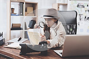 Private detective agency. Little girl is sitting at desk looking at photos with magnifying glass.