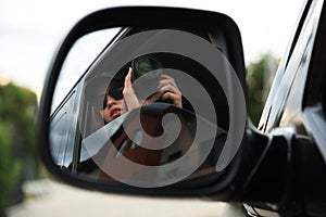 Private detective with camera spying from auto, view through car side mirror