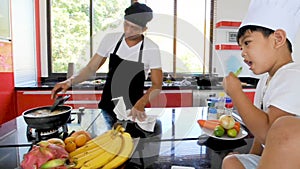 Private Thai chef cooking, his little son in chef`s hat nearby sitting on the table in a modern style home kitchen