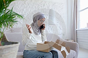 Problems with home delivery. Young muslim woman in hijab sitting on sofa and talking on the phone, holding a box on her