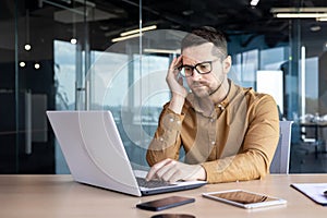 Problems at work. Pensive and serious male office worker, sitting at a desk in front of a laptop, holding his head with