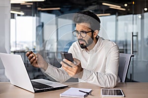 Problems with your credit card account. Worried young Indian man sitting at desk in office, holding card and phone and