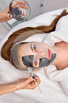 Procedure for applying black mask on face of beautiful woman, top view. Spa treatments