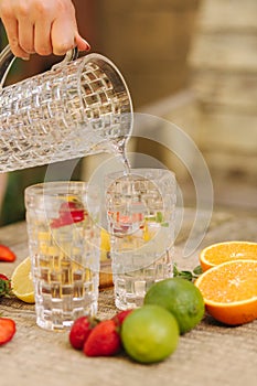 Proces of pouring cold water into a tall glass. Fresh citrus on table, orange, lemon, lime and strawberry. Food concept