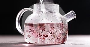 Process of brewing pink flowers tea in glass transparent teapot at dark background. Pouring hot water in kettle with tea