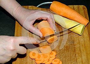 The process of cutting orange carrots with a knife and already peeled yellow and orange carrots on a light wooden cutting board
