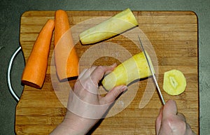 The process of cutting yellow carrots with a knife and already cleaned orange carrots on a light wooden cutting board