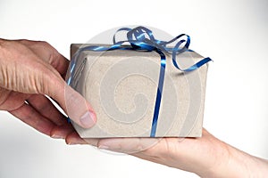 The process of giving a gift wrapped in Kraft paper with a blue ribbon on a white background. men`s and women`s hands in the fra