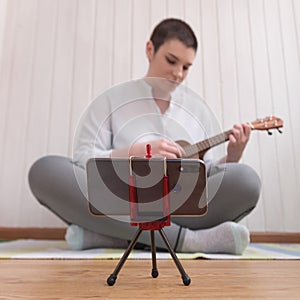 The process of learning to play the guitar online. A brunette girl is sitting with a ukulele in front of a smartphone. Teacher or