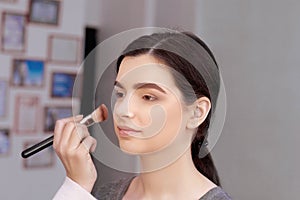 Process of making makeup. Make-up artist working with brush on model face. Portrait of young woman in beauty saloon interior.