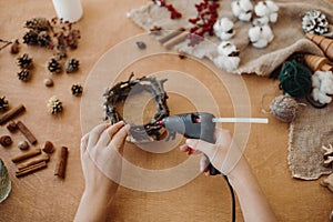 Process of making rustic christmas candle. Hands holding glue gun and making rustic festive decoration from branches, pine cones