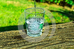 Process of pouring pure clear water into a glass from top, wooden log, green grass in the background, outdoors, health, hydration