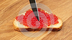Process of spreading with a knife red chum salmon caviar on a piece of bread made of white wheat flour on a wooden table