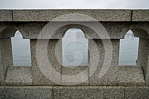 Processed gray granite stone parapet with black grainy inclusions consisting of masonry, columns and arches, water surface behind