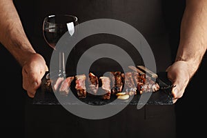 Man holding juicy grilled beef steak with spices and red wine glass on a stone cutting board on a black background