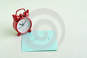 Procrastination, postpone or laziness concept, red alarm clock on sticky note with handwriting the word Later on office table photo