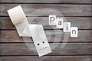 Proctology concept with pain text, toilet paper roll and painted face on wooden background top view