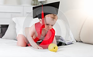 Prodigy baby in graduation cap and ribbon reaching for apple