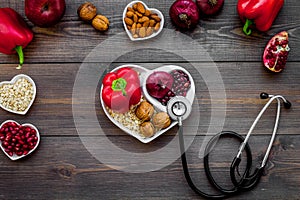 Products good for heart and blood vessels. Vegetables, fruits, nuts in heart shaped bowl near stethoscope on dark wooden