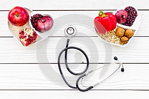 Products good for heart and blood vessels. Vegetables, fruits, nuts in heart shaped bowl near stethoscope on white