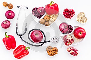 Products good for heart and blood vessels. Vegetables, fruits, nuts in heart shaped bowl near stethoscope on white