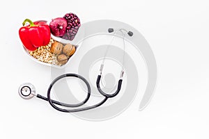 Products good for heart and blood vessels. Vegetables, fruits, nuts in heart shaped bowl near stethoscope on white