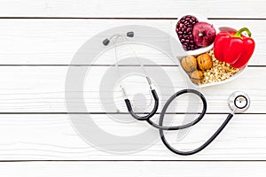 Products good for heart and blood vessels. Vegetables, fruits, nuts in heart shaped bowl near stethoscope on white