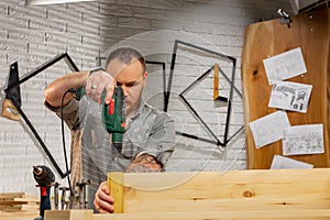 Profession, carpentry, woodwork and people concept - carpenter with electric drill drilling wood plank at workshop