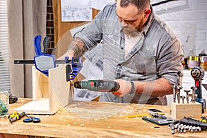 Profession, carpentry, woodwork and people concept - carpenter with electric drill drilling wood plank at workshop