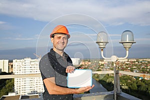 Builder in a protective helmet with a tablet for writing in his hand is standing on the roof of a building overlooking the city