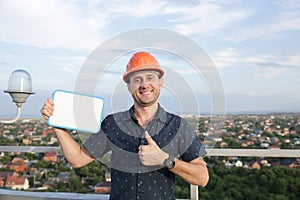 Builder in a protective helmet with a tablet for writing in his hand is standing on the roof of a building overlooking the city