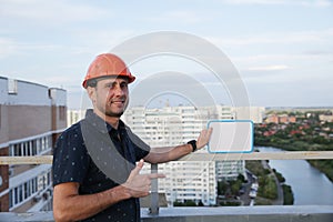 Builder in a protective helmet with a tablet for writing in his hand is standing on the roof of a building overlooking the city